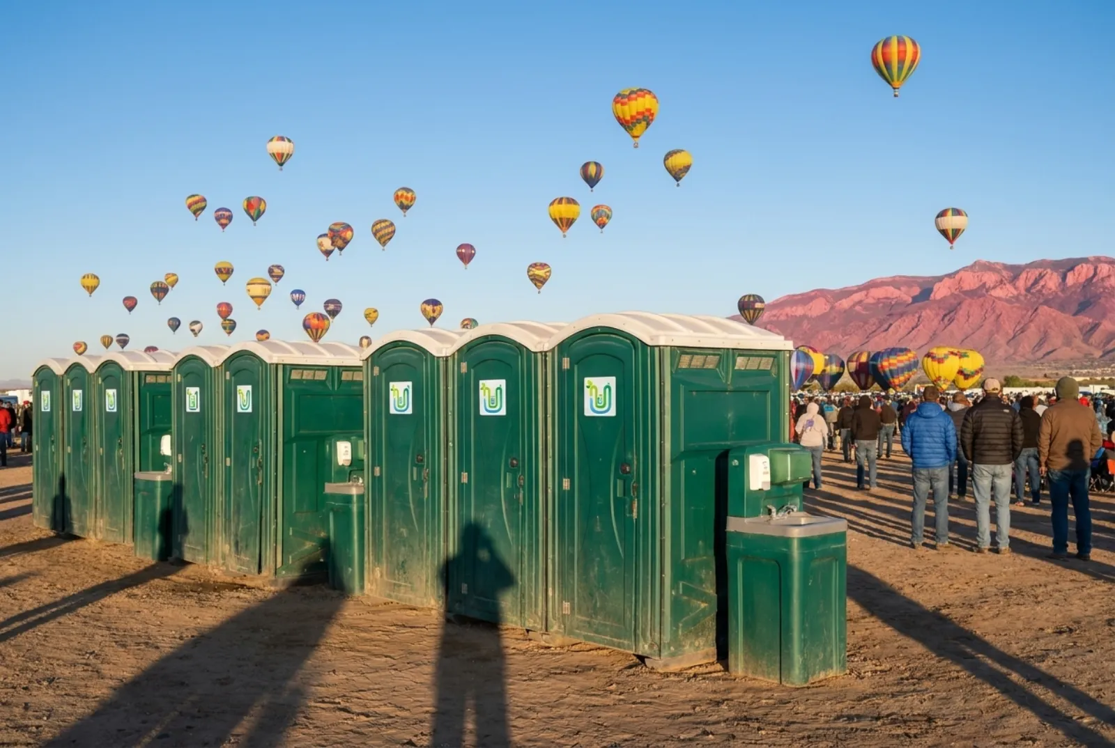 A row of portable toilets and hand-wash stations at the edge of a launch field at dawn, with colorful hot-air balloons rising and the Sandia Mountains behind