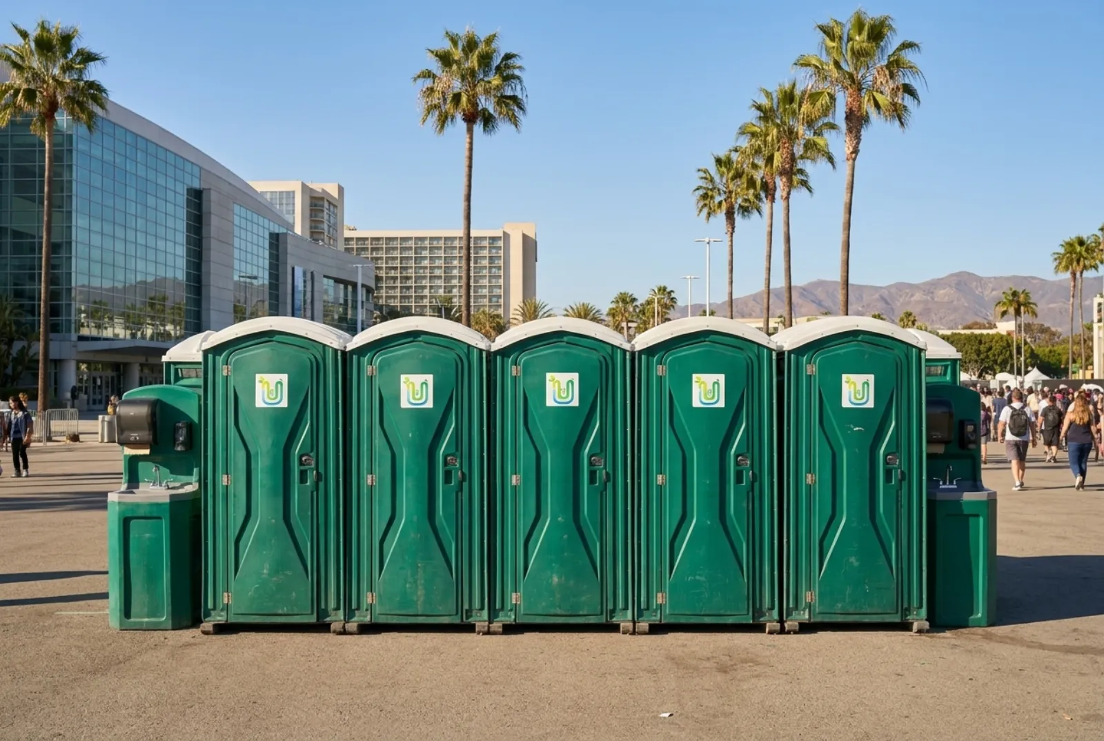 A row of green portable toilets and hand-wash stations on a paved lot lined with palm trees, with a large convention hall, hotels, and distant brown hills under a clear sky