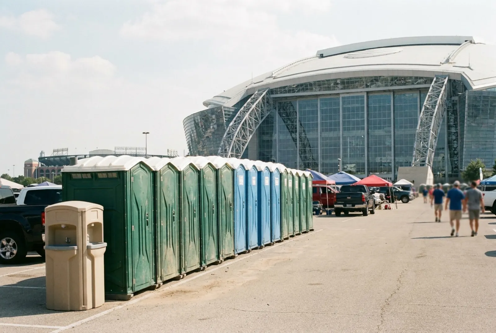 A row of portable toilets and two hand-wash stations at the edge of a large stadium parking lot in Arlington, Texas, with a retractable-roof stadium and tailgate tents behind on a hot afternoon