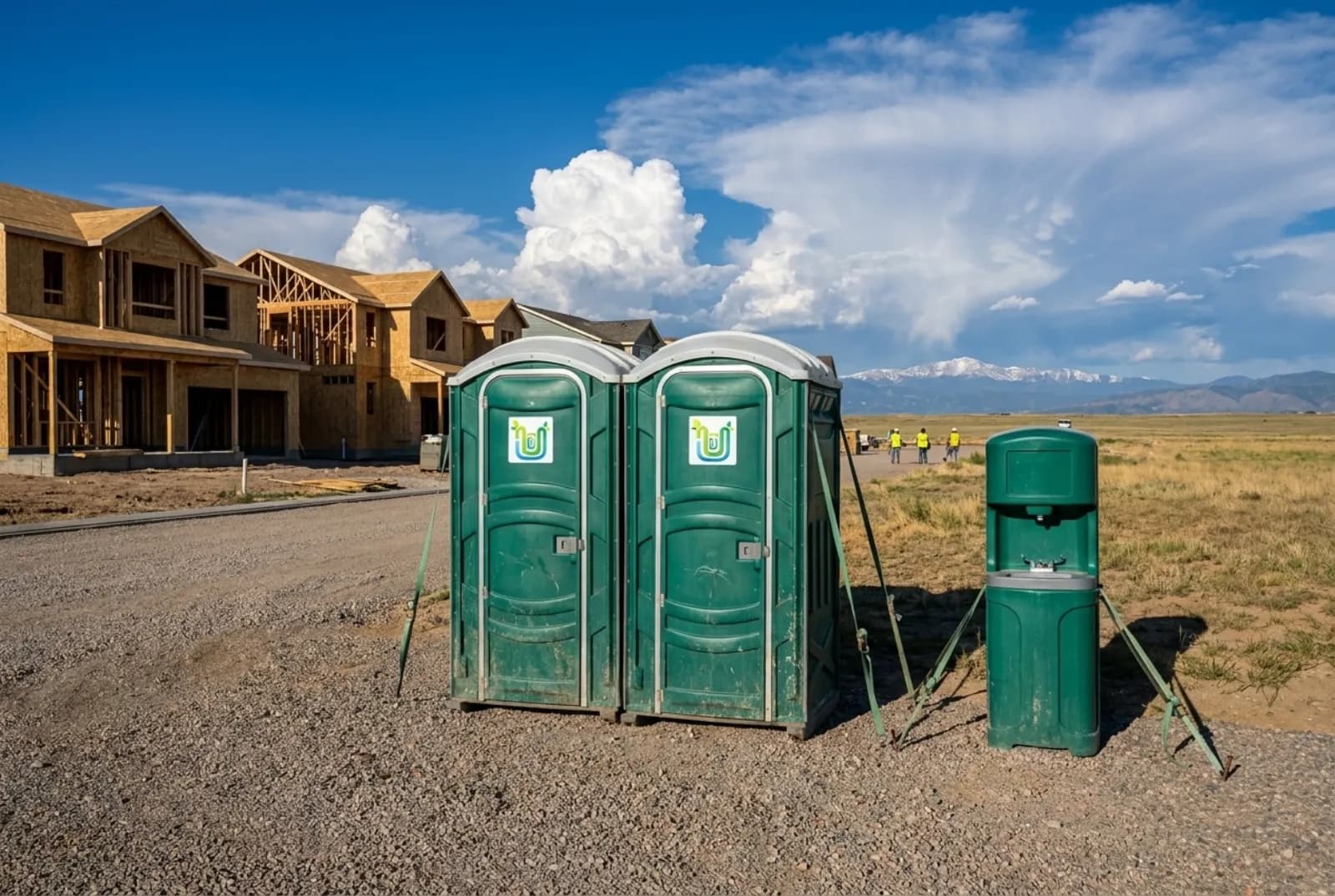 Two portable toilets with anchor straps and a hand-wash station on level gravel at a subdivision under construction on flat prairie, with storm clouds building over distant mountains