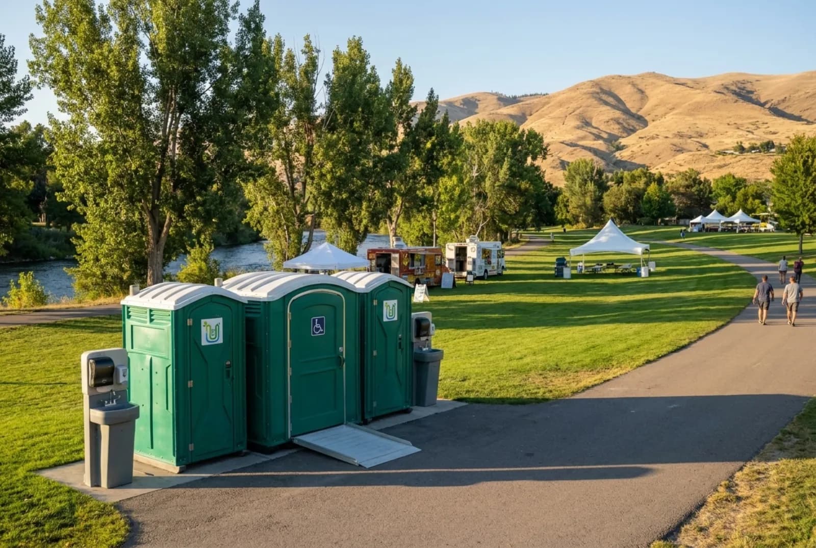 A row of portable toilets including a wheelchair-accessible unit, plus two hand-wash stations, on a paved path at the edge of a grassy riverside park with cottonwood trees, event tents, food trucks, and dry tan foothills behind
