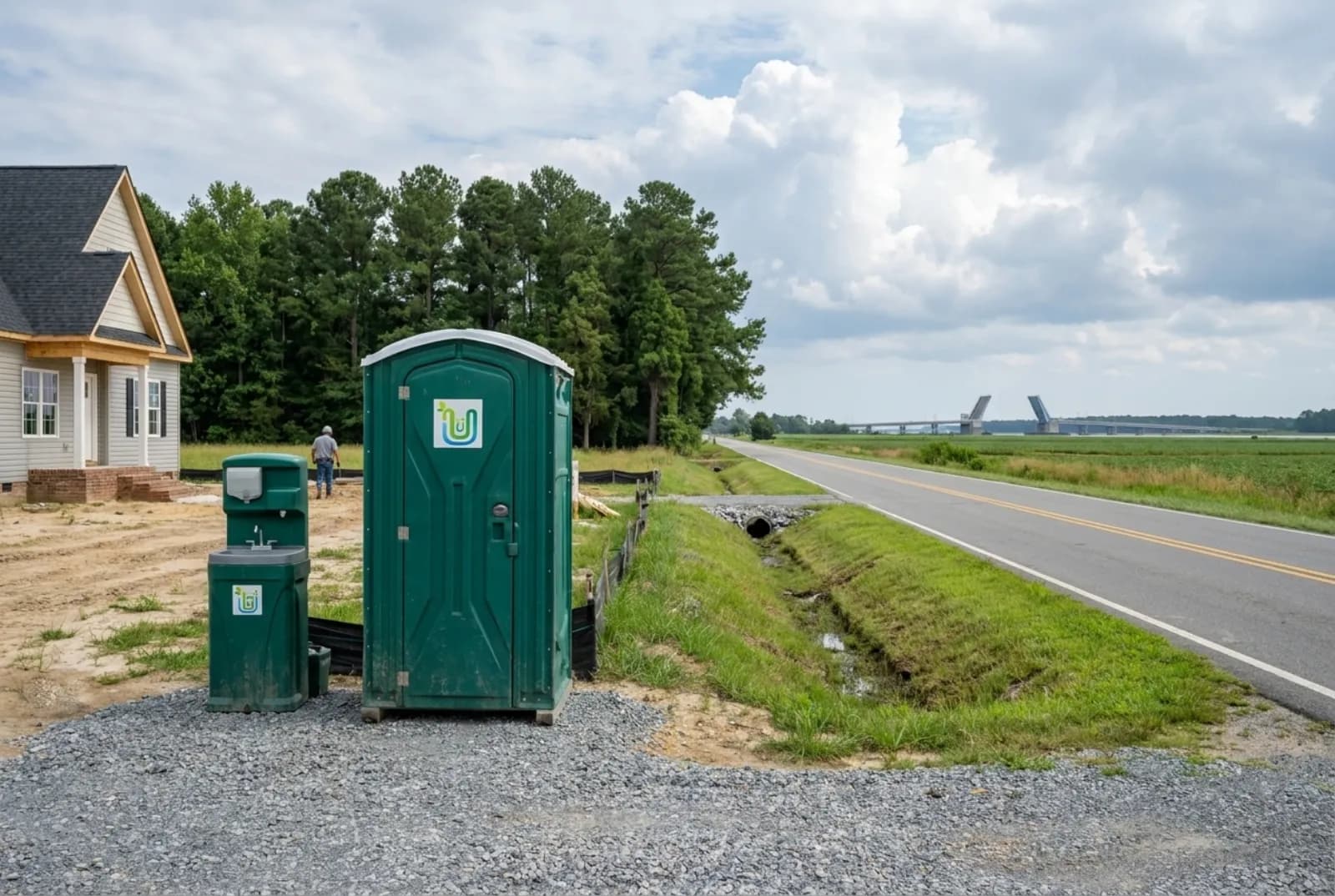 A portable toilet and hand-wash station on a gravel pad at a new-home site in Chesapeake, set back from a grassy roadside ditch, with farm fields and a distant drawbridge.