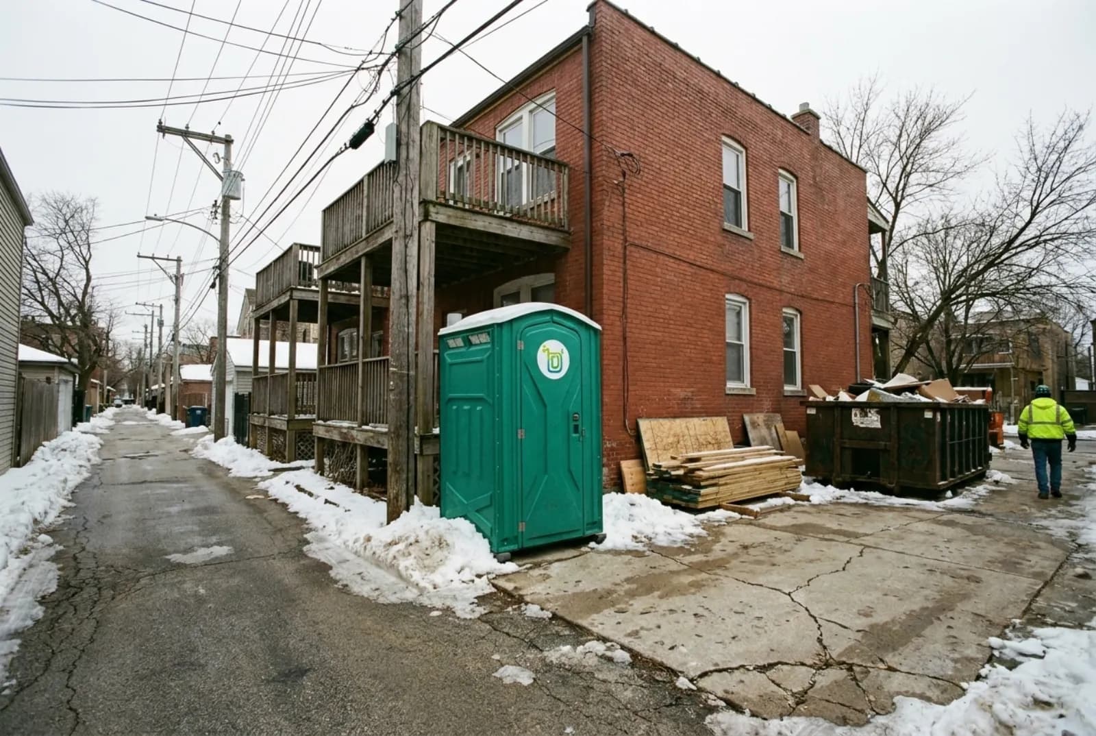 A portable toilet on level concrete behind a brick two-flat under renovation, beside a snowy paved alley lined with wooden back porches and garages under a gray winter sky