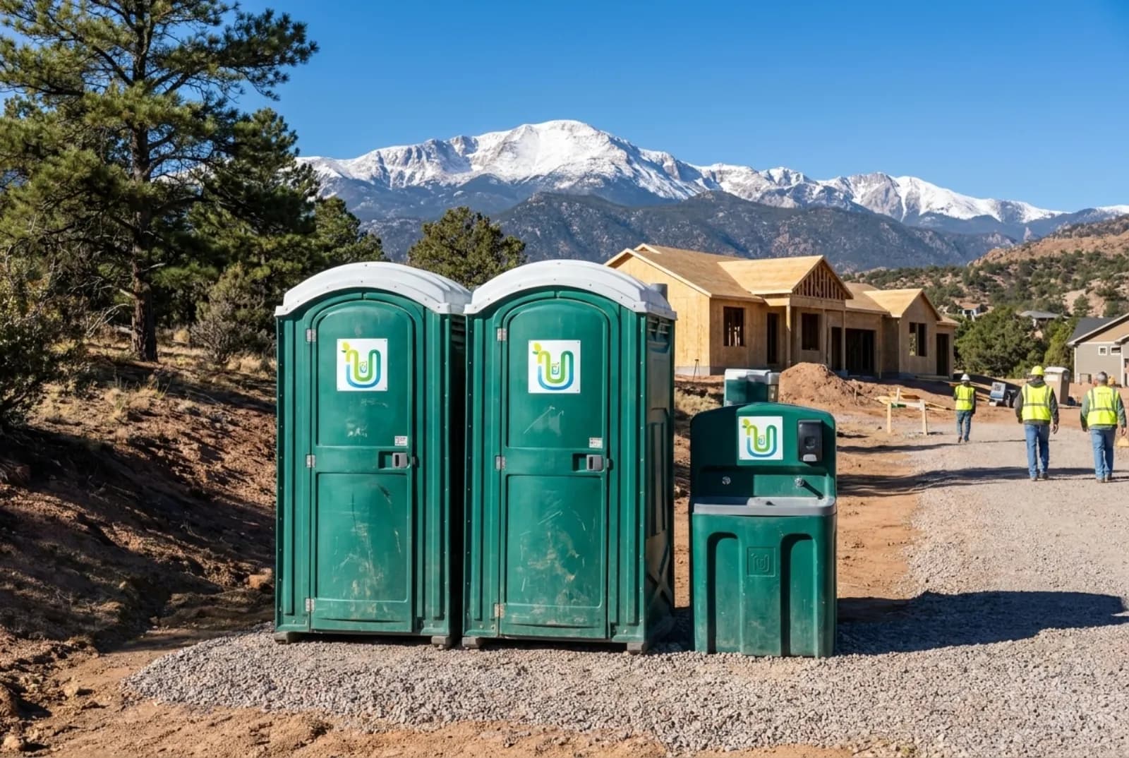 Two portable toilets and a hand-wash station staked on level gravel at a foothills home construction site in Colorado Springs, with Pikes Peak and ponderosa pines behind under a clear blue sky