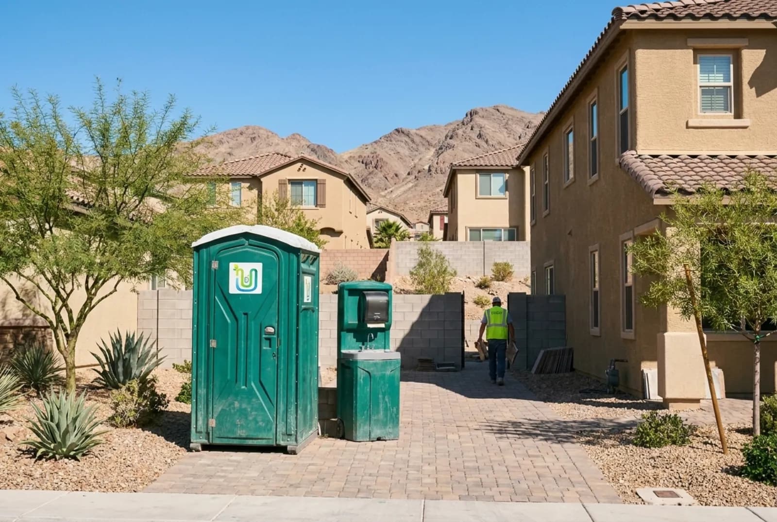 A portable toilet and a hand-wash station in the shaded side yard of a stucco home under remodel in a Henderson, Nevada master-planned neighborhood, with desert landscaping and brown foothills behind