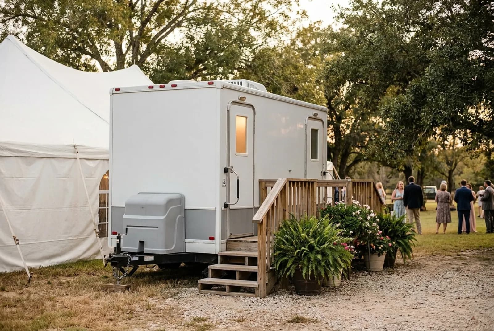 A white two-stall luxury restroom trailer with wooden steps and potted plants beside a wedding tent at a rural venue outside Houston