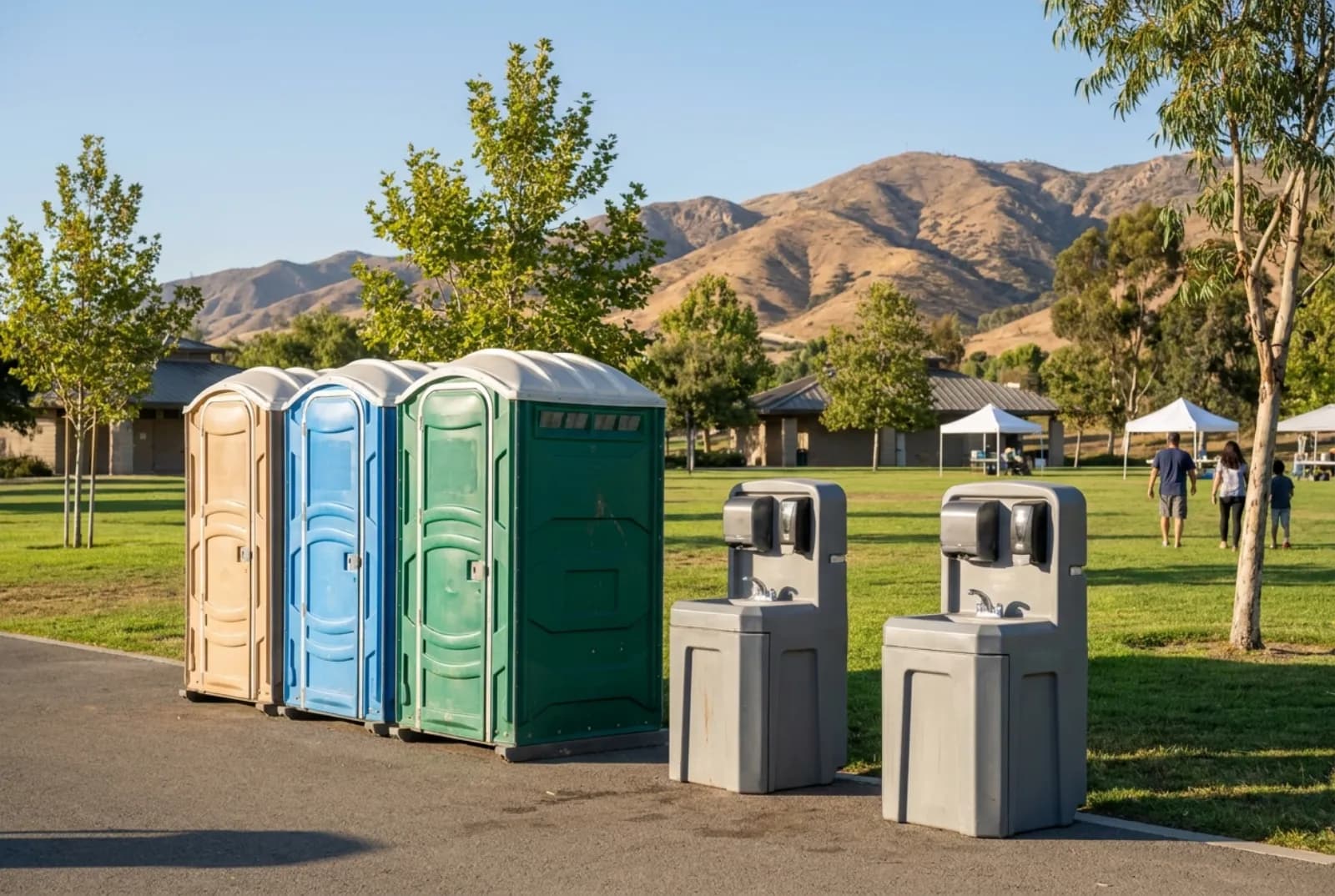 A tidy row of portable toilets and two hand-wash stations on a paved path beside a manicured park lawn, with event canopies, young trees, and dry golden foothills behind