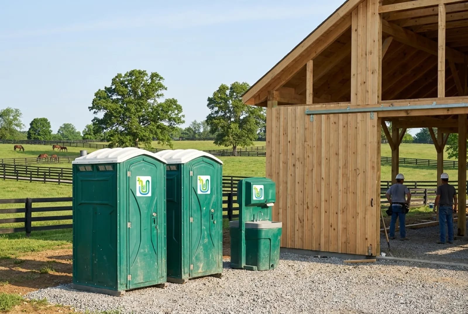 Two portable toilets and a hand-wash station on gravel beside a horse barn under construction, with black plank fences, rolling green pasture, and grazing horses behind