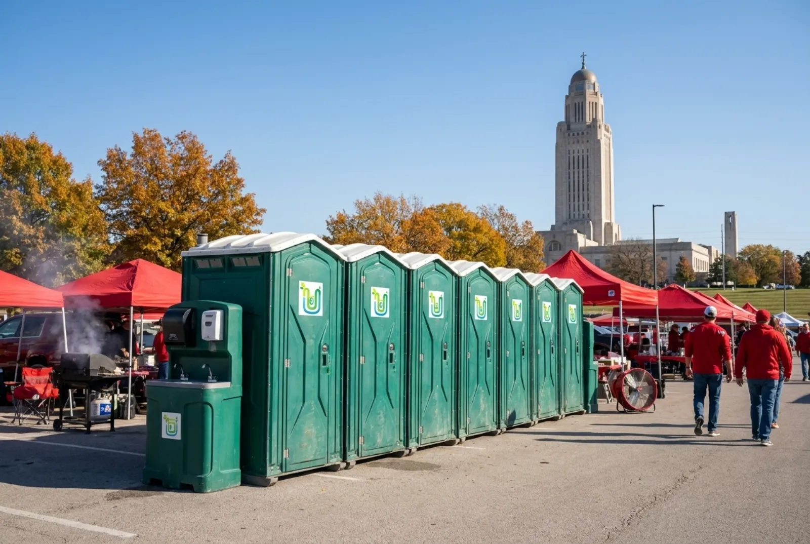 A long bank of portable toilets with hand-wash stations on asphalt at the edge of a tailgate lot with red tents, fall trees, and a tall limestone capitol tower on the skyline