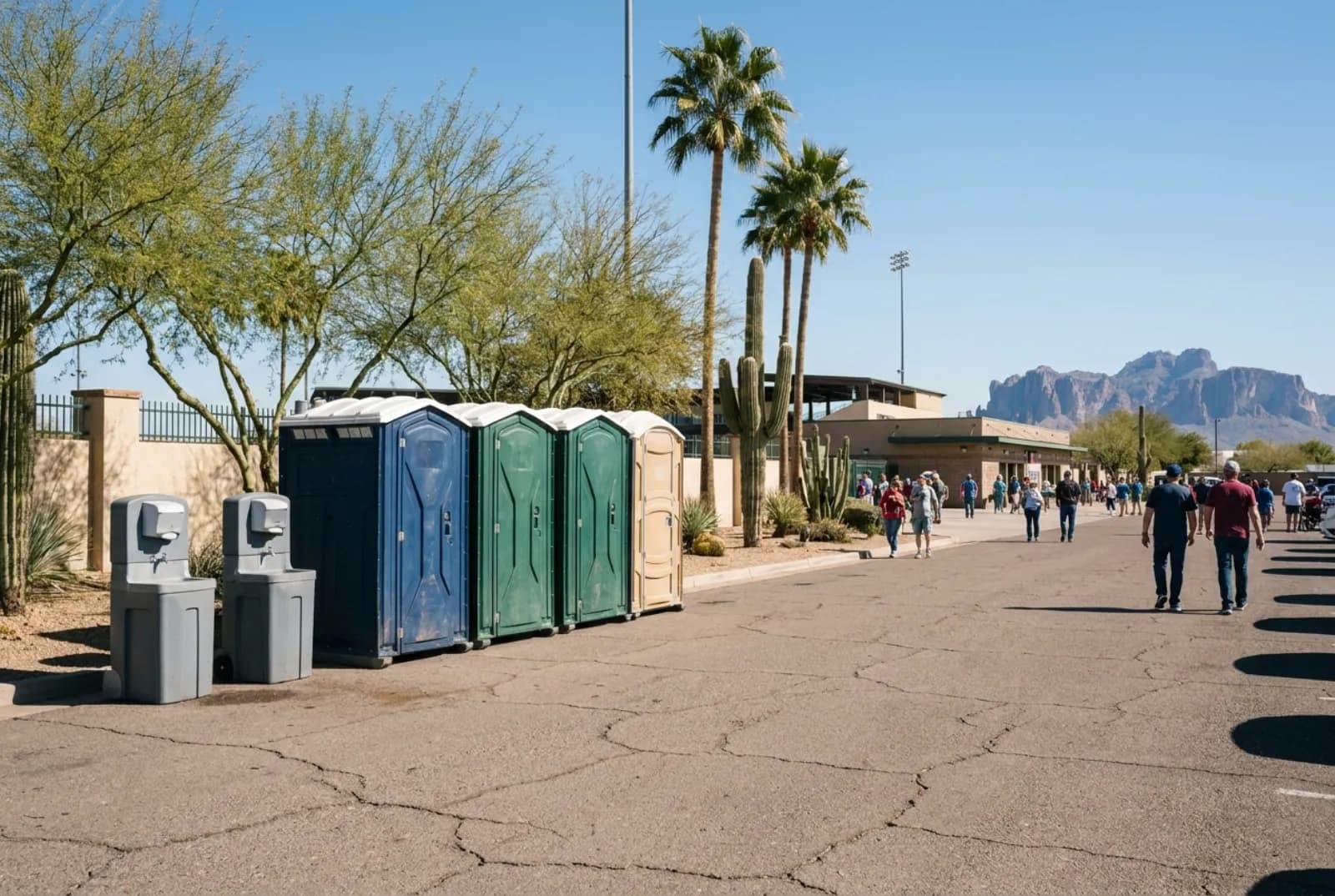 A bank of portable toilets and two hand-wash stations on asphalt at the edge of a ballpark parking lot, with palm trees, desert plants, and rugged mountains under a clear sky