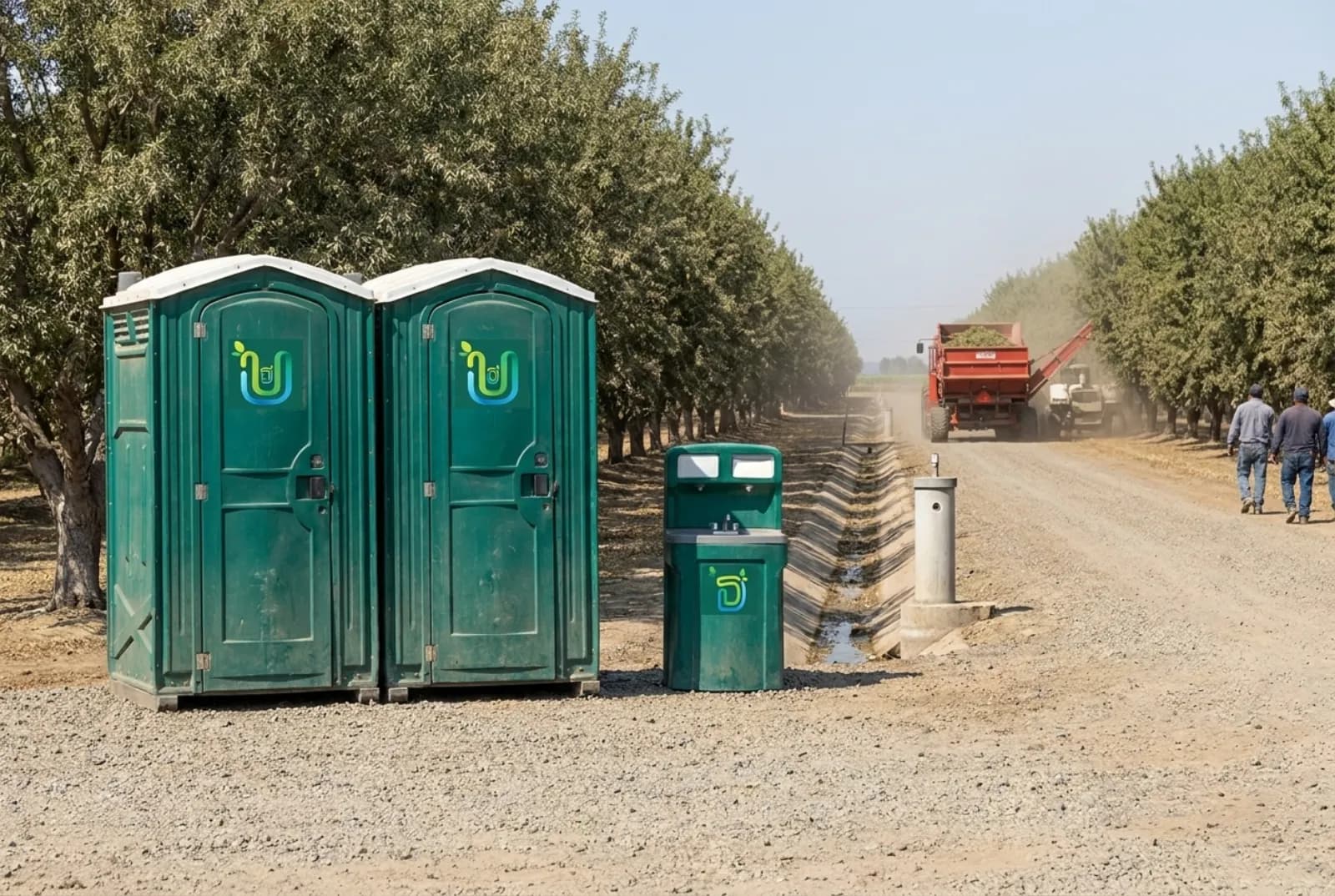 Two portable toilets and a hand-wash station on a gravel lane at the edge of an almond orchard, with harvest equipment working the rows and an irrigation ditch beside the lane