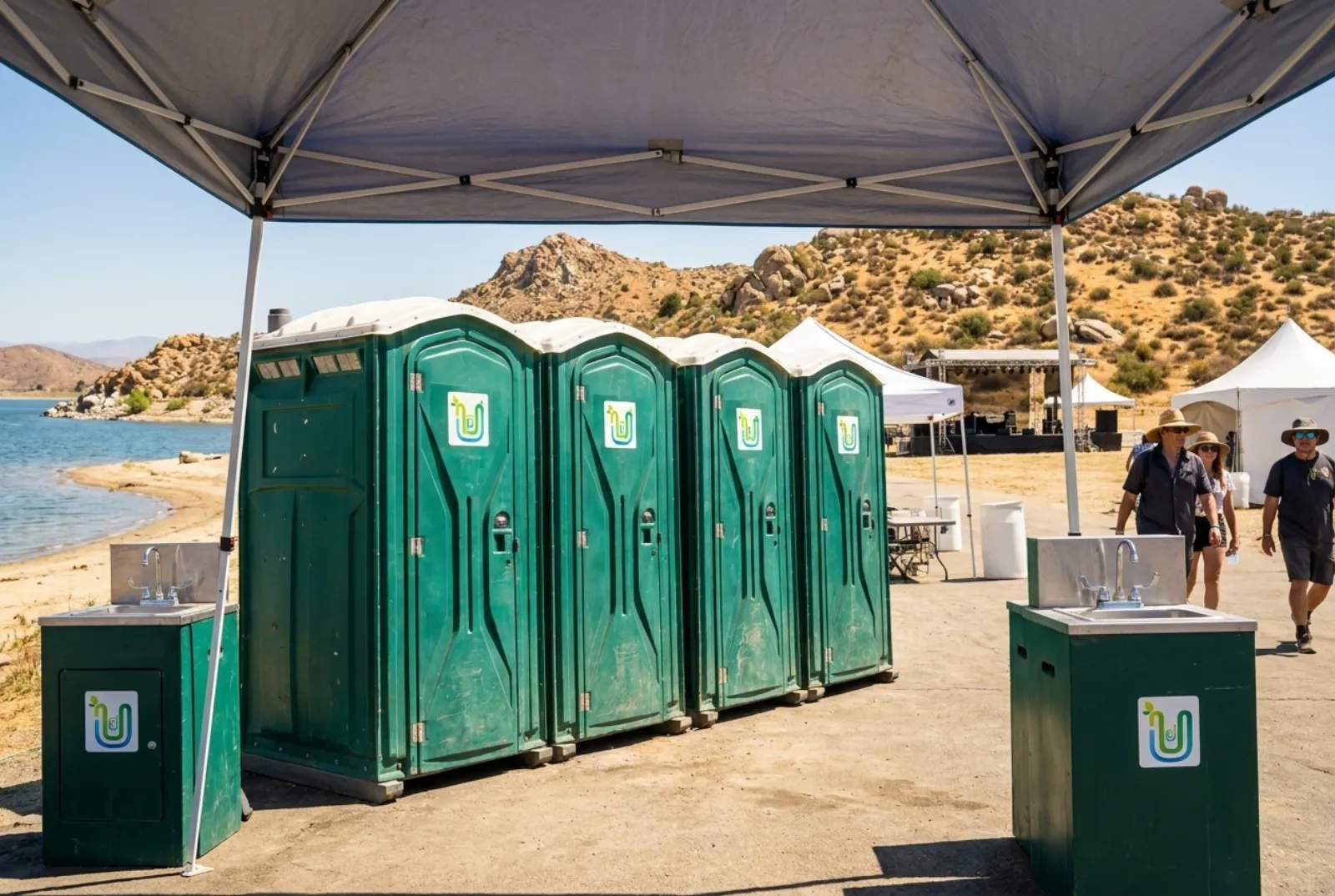 A row of portable toilets and two hand-wash stations under a shade canopy at an outdoor summer event beside a lake, with dry boulder-covered hills and white event tents behind