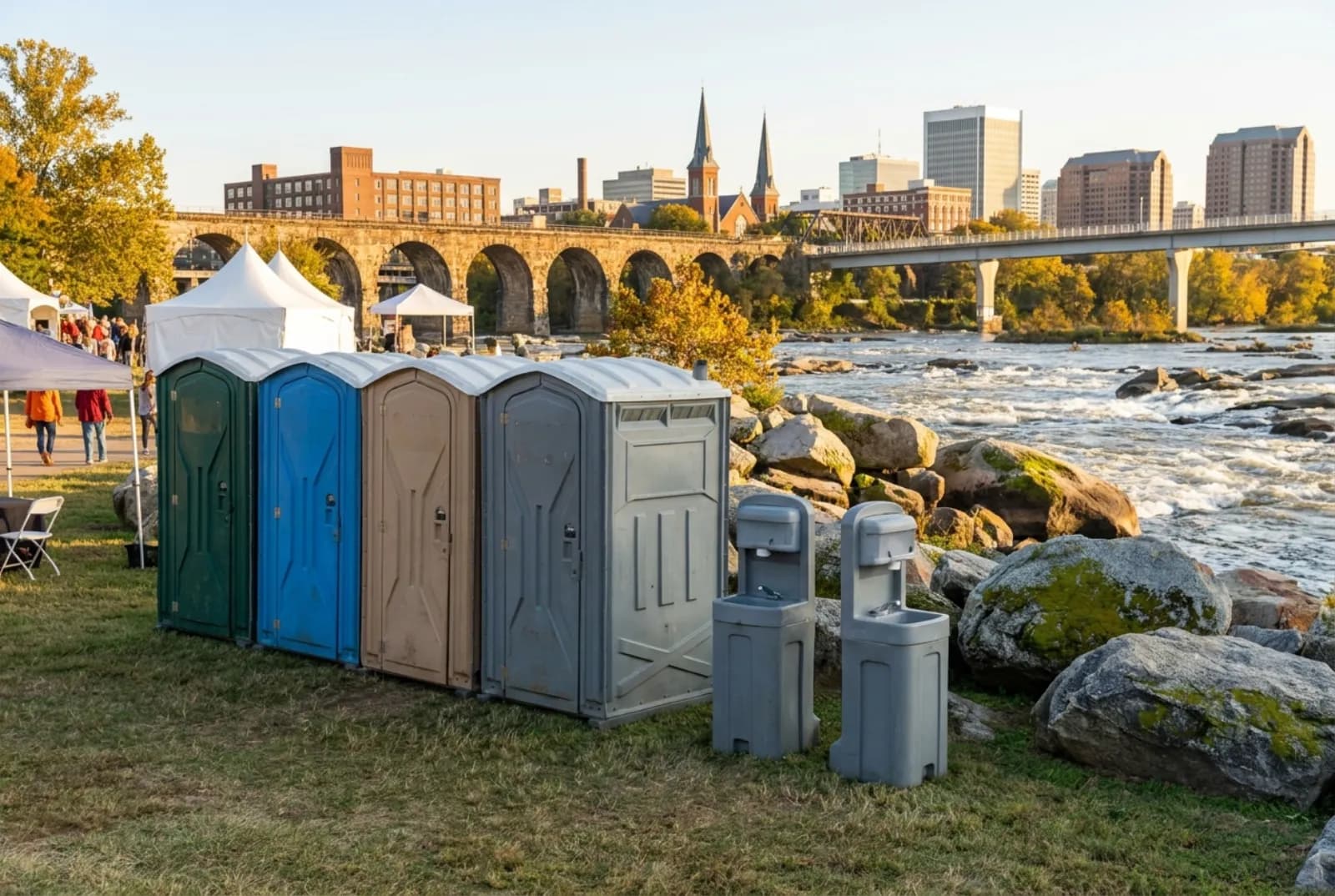 A row of portable toilets and hand-wash stations on mowed grass at a riverside festival, with the rocky James River rapids, a stone trestle, and the downtown Richmond skyline behind.