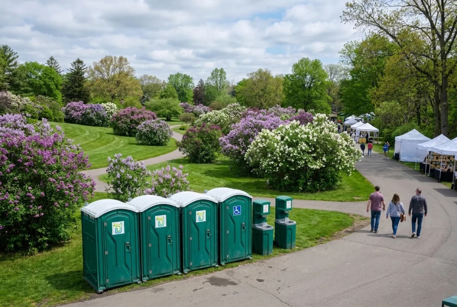 A row of portable toilets, a wider accessible unit, and two hand-wash stations on a paved park road beside blooming lilac shrubs and festival tents in a hillside park in spring.