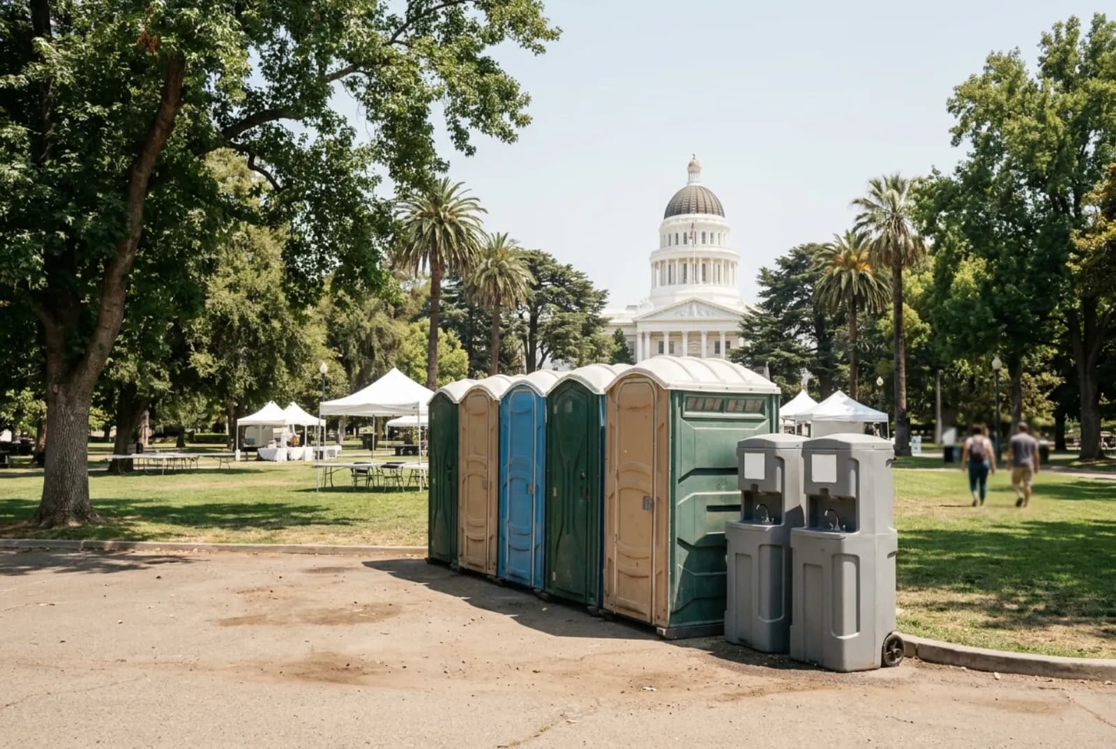 A row of portable toilets and two hand-wash stations on pavement beside a tree-shaded lawn with white event canopies, with the white California State Capitol dome behind