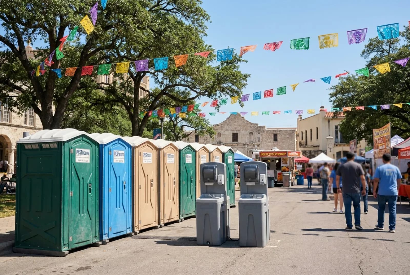 A row of portable toilets and two hand-wash stations on pavement at the edge of a festival plaza under colorful paper banners, with live oaks and stucco buildings behind