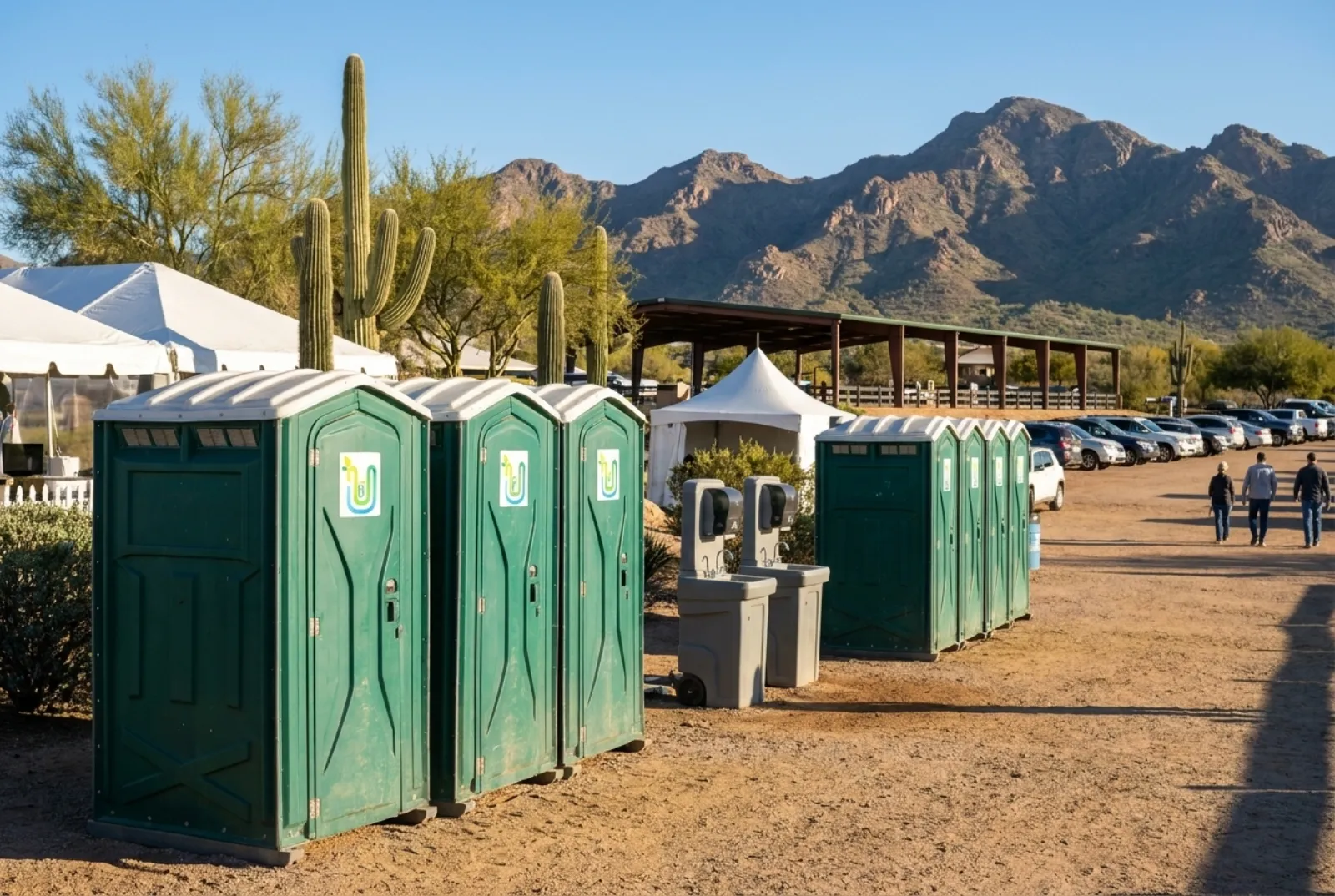 A row of portable toilets and two hand-wash stations on level desert ground at the edge of an event grounds with white tents, a covered arena, parked cars, saguaros, and the McDowell Mountains behind
