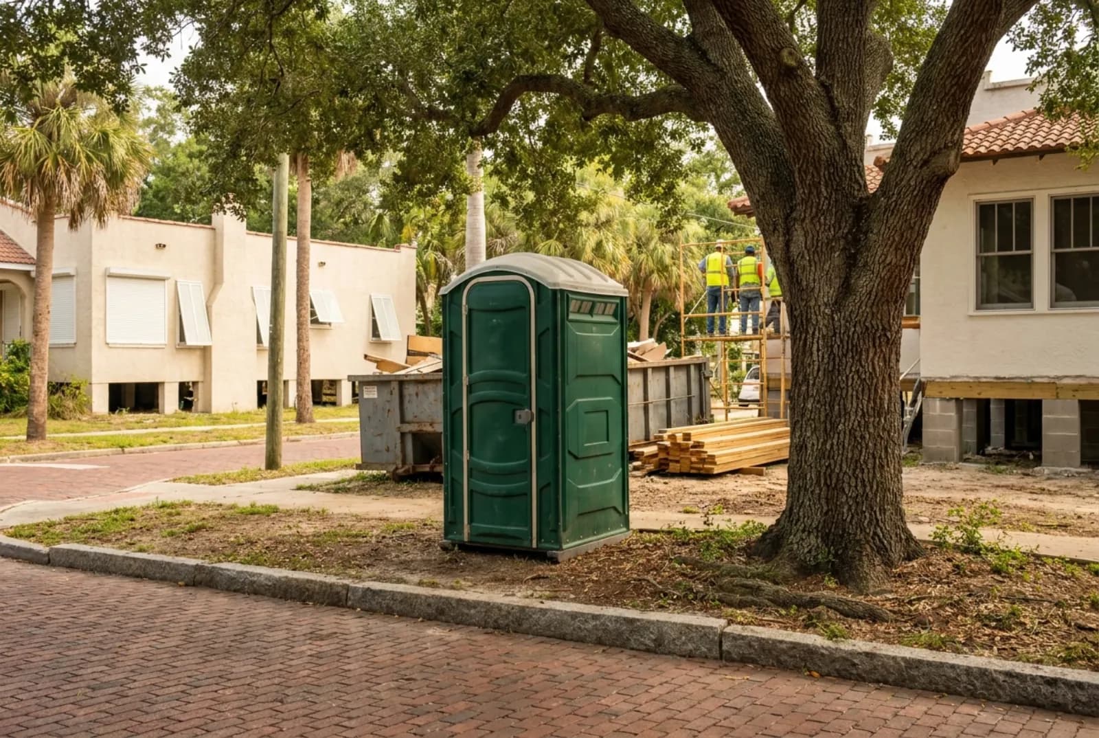 A portable toilet in the shaded side yard of a 1920s St. Petersburg bungalow being raised on new piers, with a red-brick street and granite curb in front.