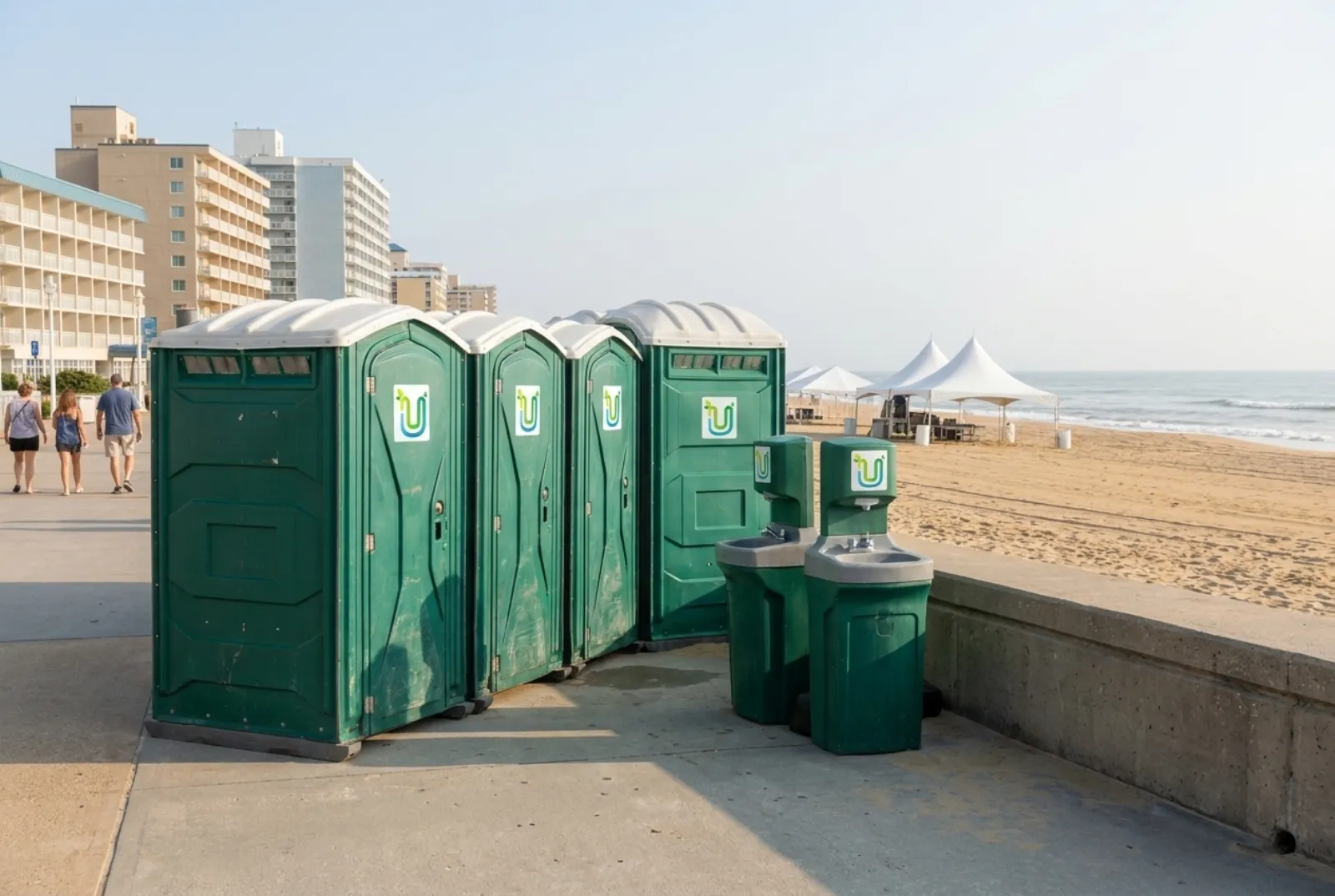 A row of portable toilets and two hand-wash stations on pavement beside a concrete oceanfront boardwalk, with a wide sandy beach, surf, white tents, and a line of hotels behind