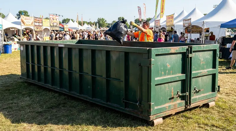 Roll-off dumpster at a festival grounds with a worker tossing a trash bag